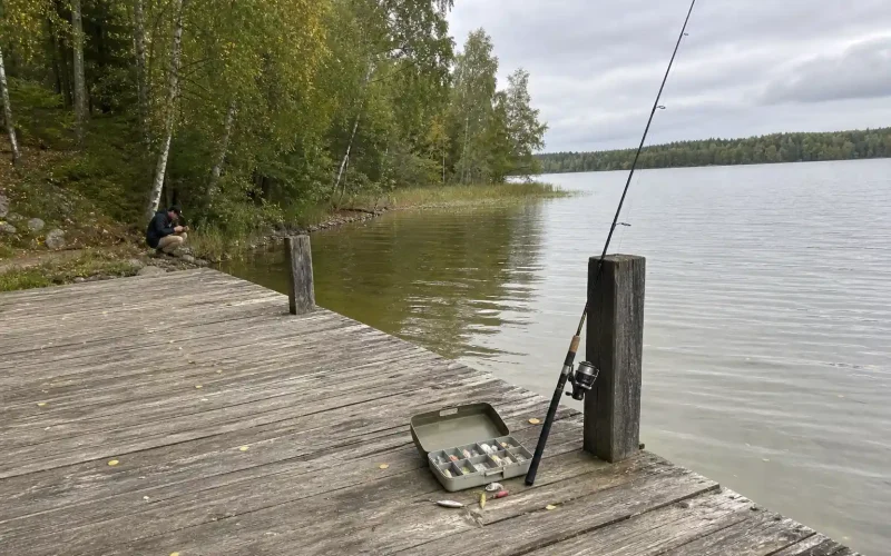 Öppen betesask och spinnspö på brygga vid sjö, person knyter fiskelina i bakgrunden. Vilka köder vi använder i september 2...
